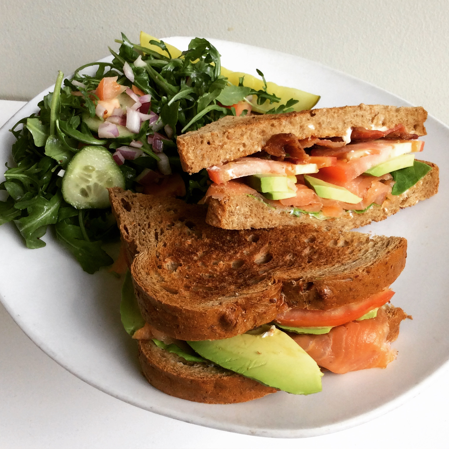 Sandwich with smoked salmon, avocado, bacon, and aioli, and a side salad, on a white plate
