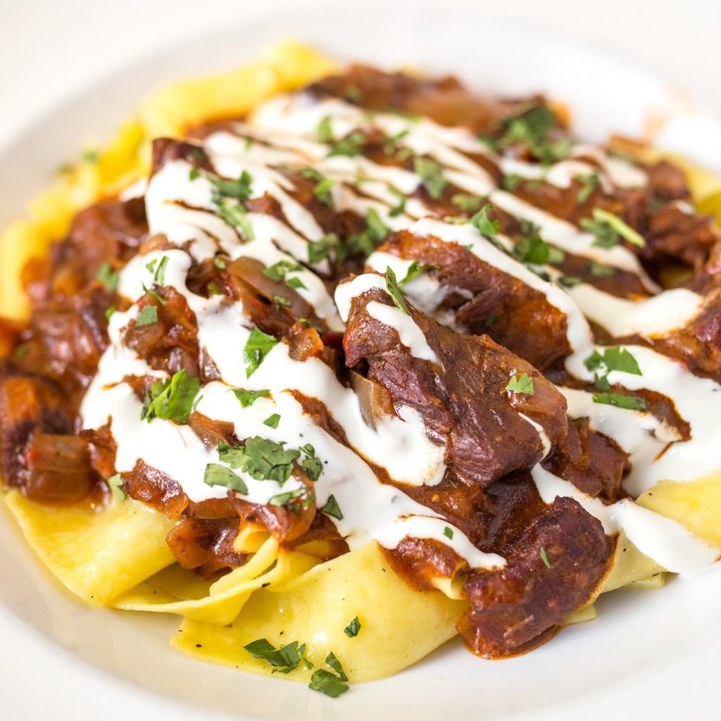 Bowl of beef goulash over pappardelle pasta and drizzled with sour cream