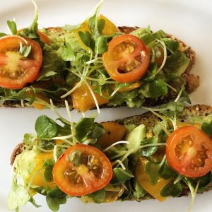white plate with baguette slices topped with mashed avocado, yellow and red tomatoes, microgreens
